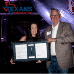 Query successful Man and woman hold a framed document with a pen, Texans for Medical Freedom logo. Aircraft in background.