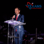 Man in a blue suit speaking at a clear podium with a microphone, Texans for Medical Freedom logo above.