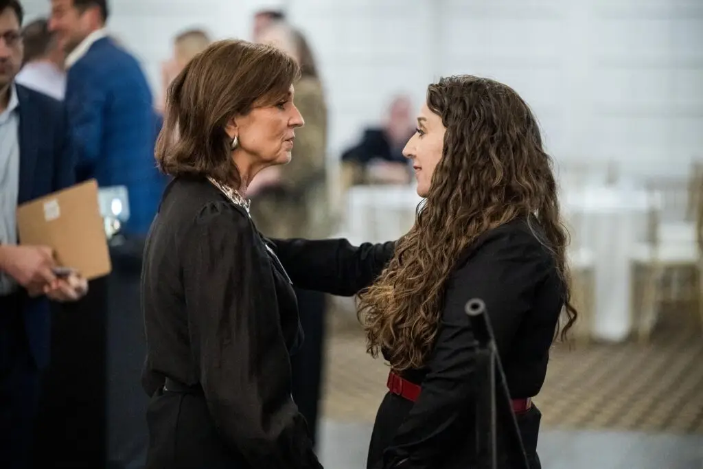 An eye-level shot of two women talking intently at an indoor event. The woman on the left has short brown hair and is wearing a dark suit jacket and large hoop earrings. The woman on the right has long, curly brown hair, is wearing a black outfit with a red belt, and has her hand on the other woman's shoulder. People are blurred in the background.
