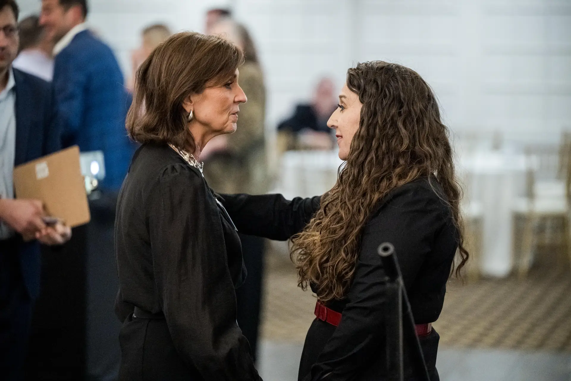 An eye-level shot of two women talking intently at an indoor event. The woman on the left has short brown hair and is wearing a dark suit jacket and large hoop earrings. The woman on the right has long, curly brown hair, is wearing a black outfit with a red belt, and has her hand on the other woman's shoulder. People are blurred in the background.