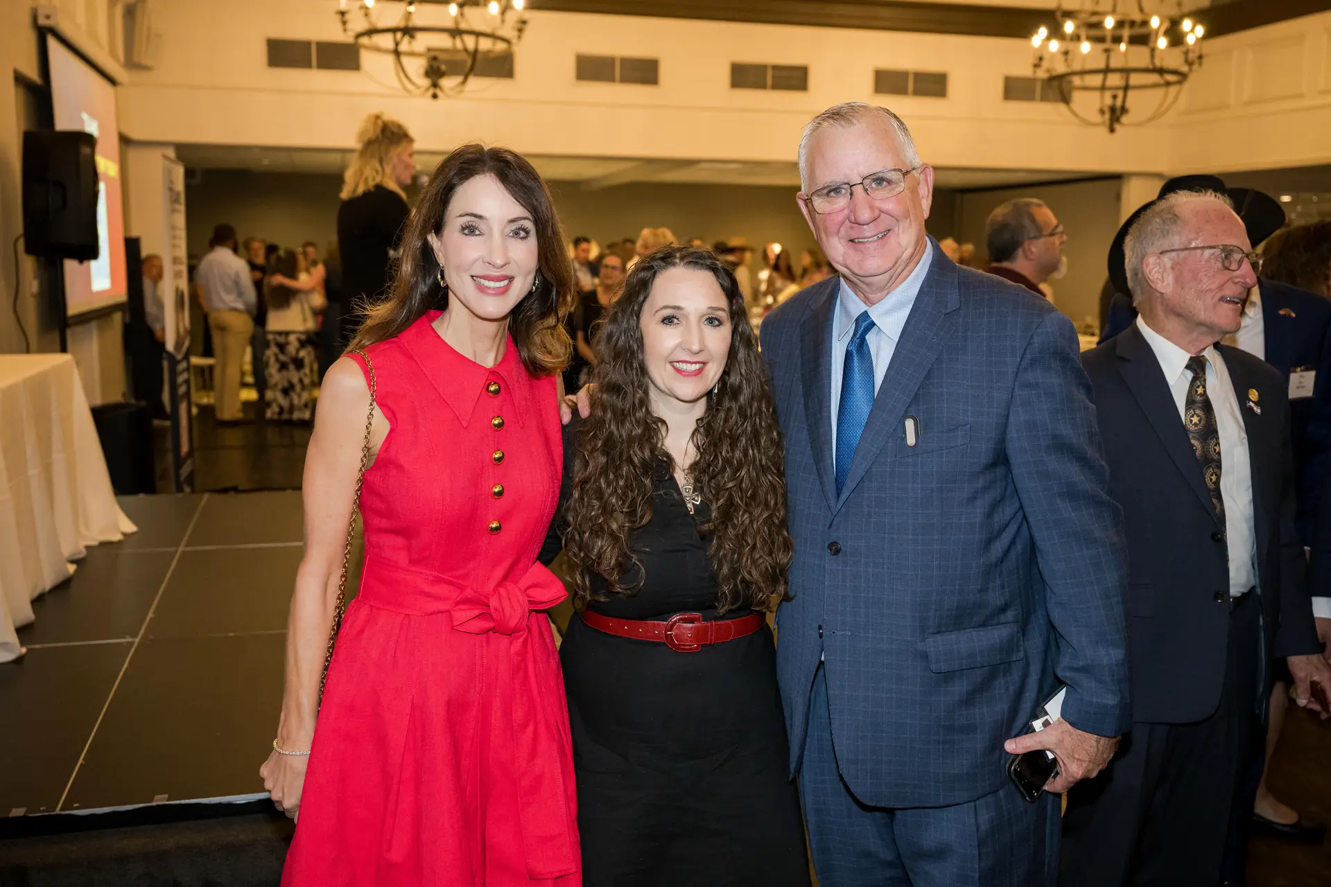 A group portrait of three smiling people at an indoor event: a woman in a bright red, sleeveless dress on the left, a woman with long, curly dark hair and a black outfit with a red belt in the center, and a man in a blue textured suit and patterned tie on the right. A crowd of people is blurred in the background.