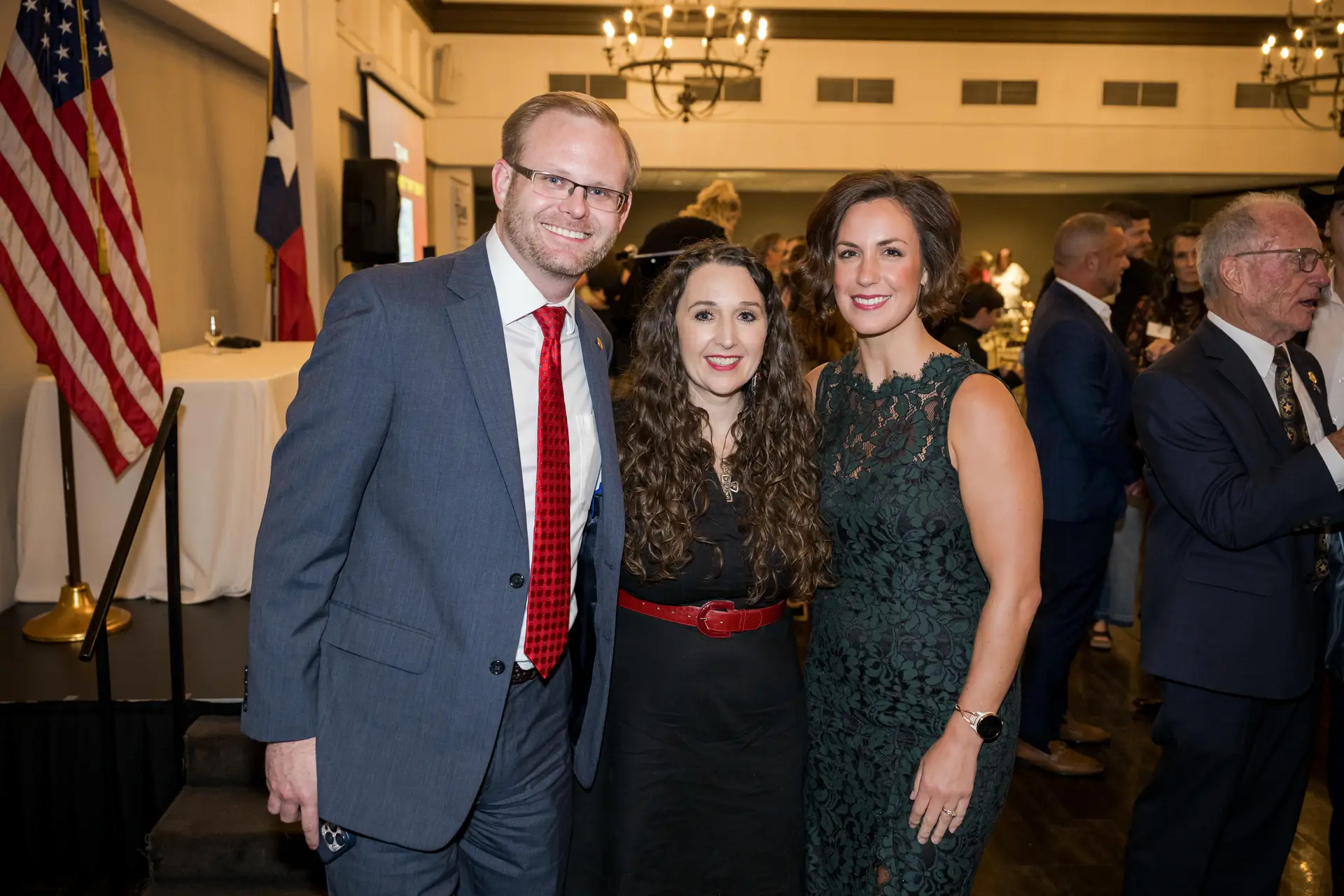 A smiling group portrait of a man in a gray suit and red tie, a woman with long, curly dark hair and a black dress with a red belt, and a woman in a dark green lace dress at a formal indoor event. The American and Texas flags are visible in the background.