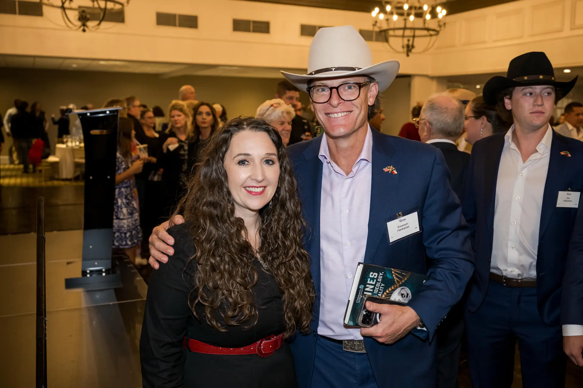 A smiling group portrait of a man in a gray suit and red tie, a woman with long, curly dark hair and a black dress with a red belt, and a woman in a dark green lace dress at a formal indoor event. The American and Texas flags are visible in the background.