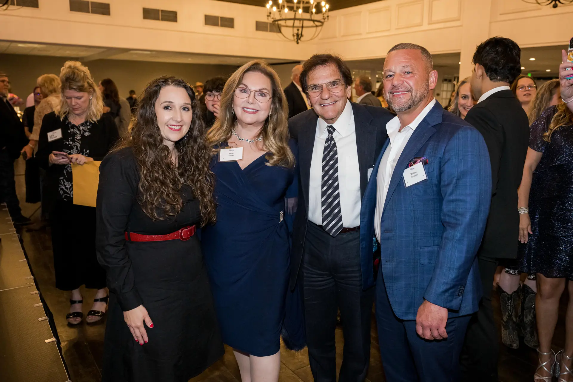 smiling group of four people posing for a photo at an indoor event: a woman with curly hair and a red belt, a woman in a navy dress and glasses, a man in a dark suit and striped tie, and a man in a blue plaid jacket. Other guests are visible in the background.