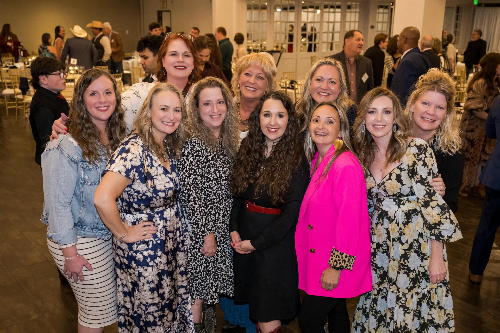 A smiling group portrait of ten women at an indoor event, standing in a crowd. The women are dressed in various styles, including a blue floral dress, a pink blazer, and a black dress with a red belt.