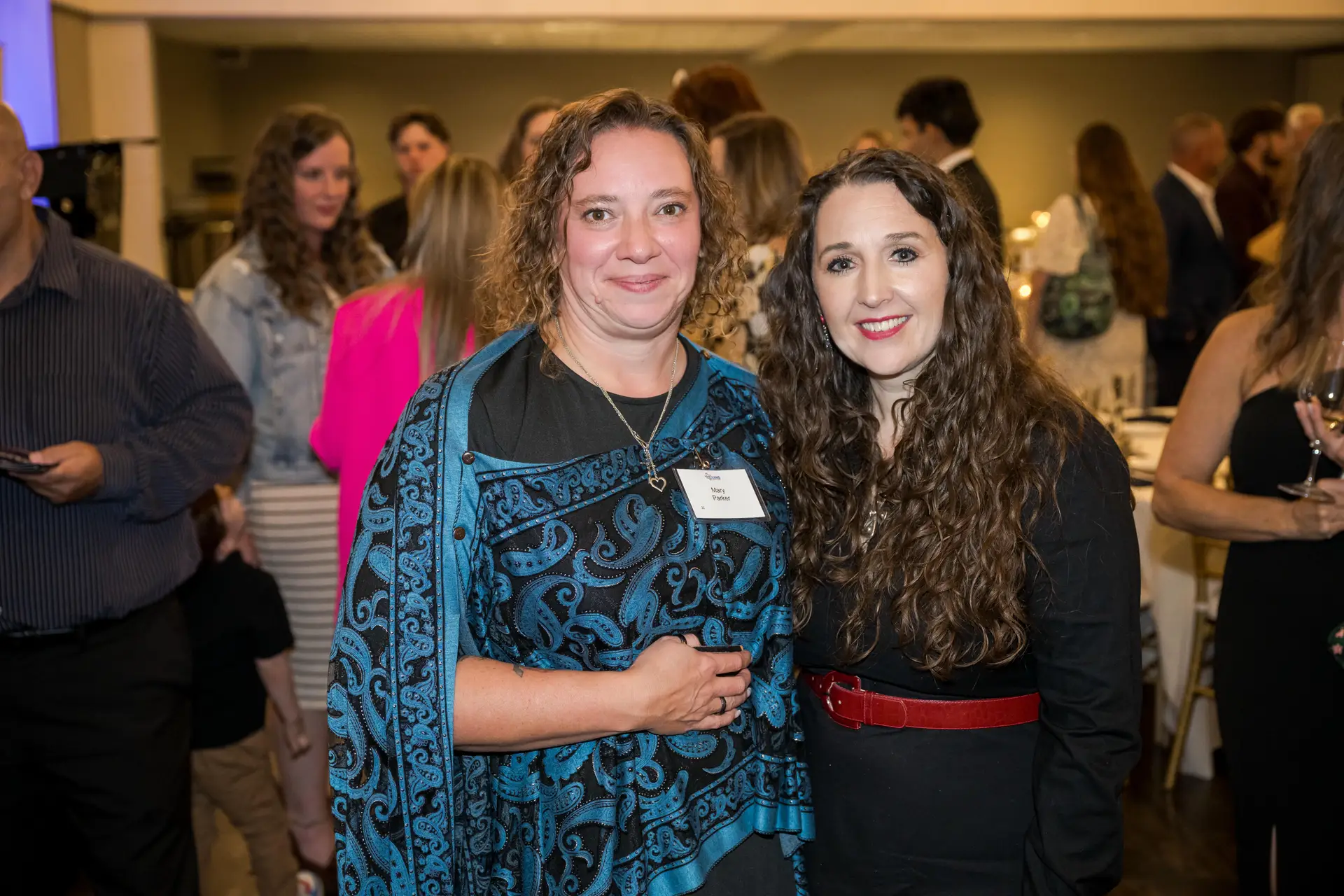 A group portrait of two smiling women at a large indoor event. The woman on the left wears a black shirt and a blue and black patterned shawl. The woman on the right has long, curly dark hair and is wearing a black dress with a red belt. Guests are visible in the blurred background.