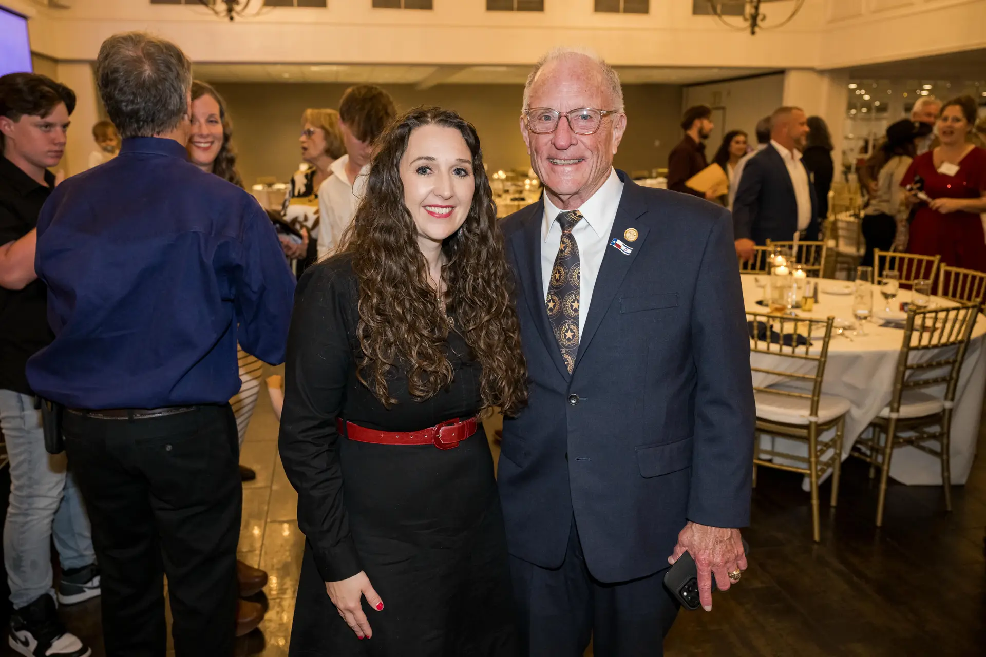 A group portrait of a smiling woman with long, curly dark hair and a red belt, standing next to an older man in a navy suit and patterned tie, at a formal indoor event. Other guests and dining tables are visible in the background.