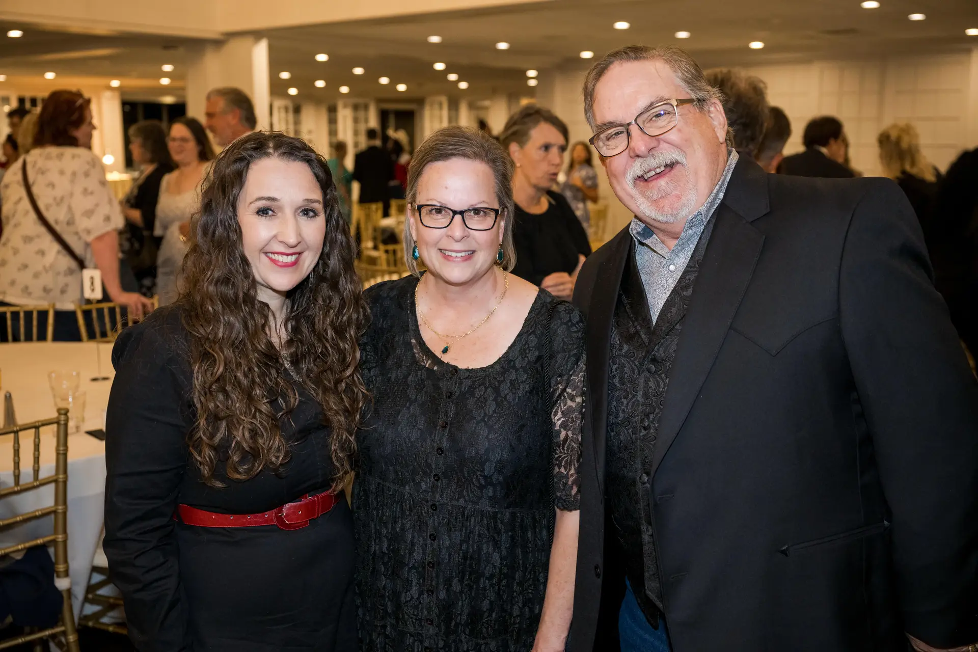 A group portrait of a smiling woman with long, curly dark hair and a red belt, a woman in a black lace dress and glasses, and a man in a black suit with a patterned vest, at a lively indoor event with many people in the background.