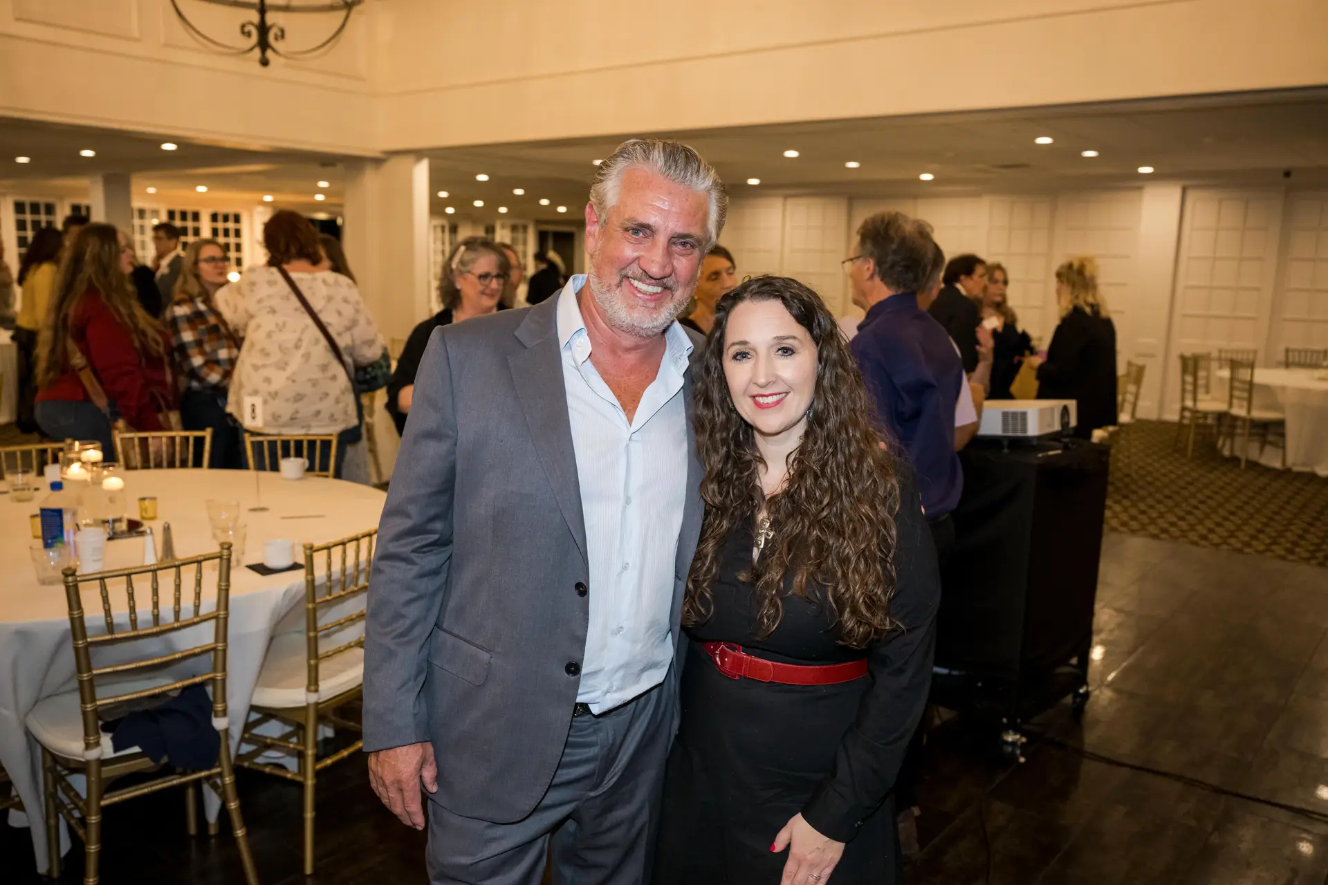 Query successful A man with gray hair and a beard, wearing a gray suit and open-collared shirt, is smiling and posing with his arm around a woman with long, curly dark hair and a red belt over a black dress. They are at an indoor event with round tables and white chairs in the background.